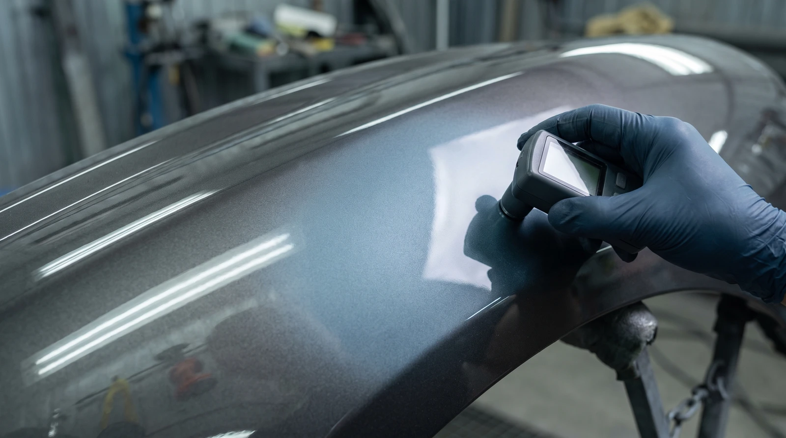 Gloved hand holding a handheld colour measuring device against a metallic grey-blue car panel with a blended paint repair